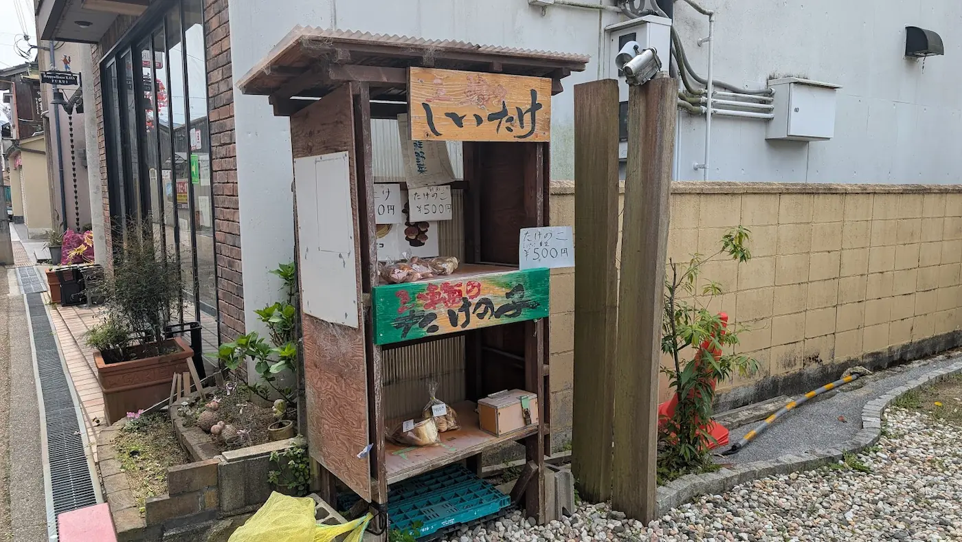 参道沿いにある、手書きの看板が並ぶ木造の無人野菜販売所。しいたけやたけのこが並んでいる。