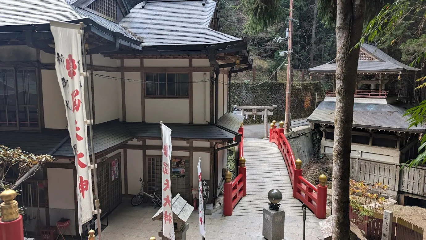 長い階段を下りきった先に見える脳天大神の境内。朱色の手すりが鮮やかな橋と、周囲に立つ白いのぼり旗、神社の建物の風景。