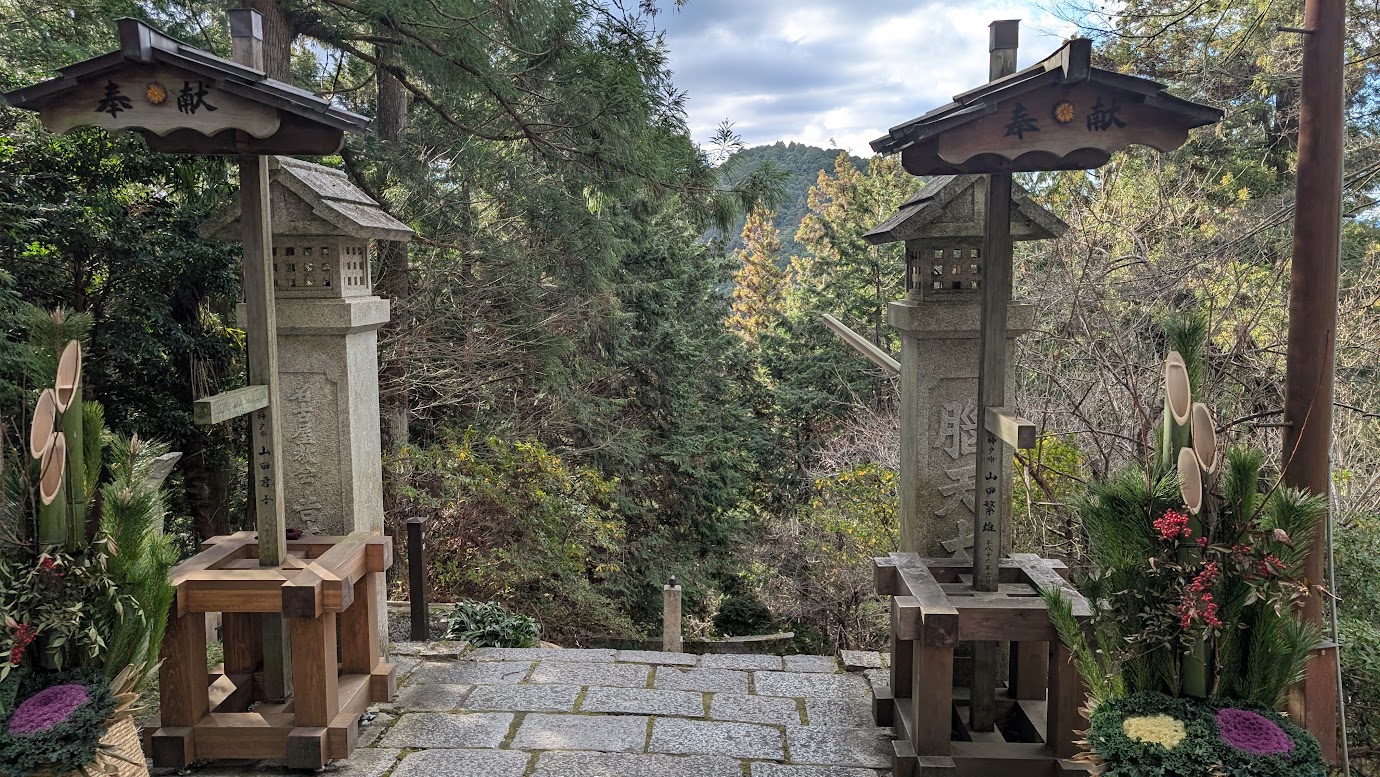 金峯山寺の境内にある「脳天大神」への入り口。正月飾りの立派な門松が供えられた石碑と石灯籠の間から、深い谷底の森へと続く石段の始まりの風景。