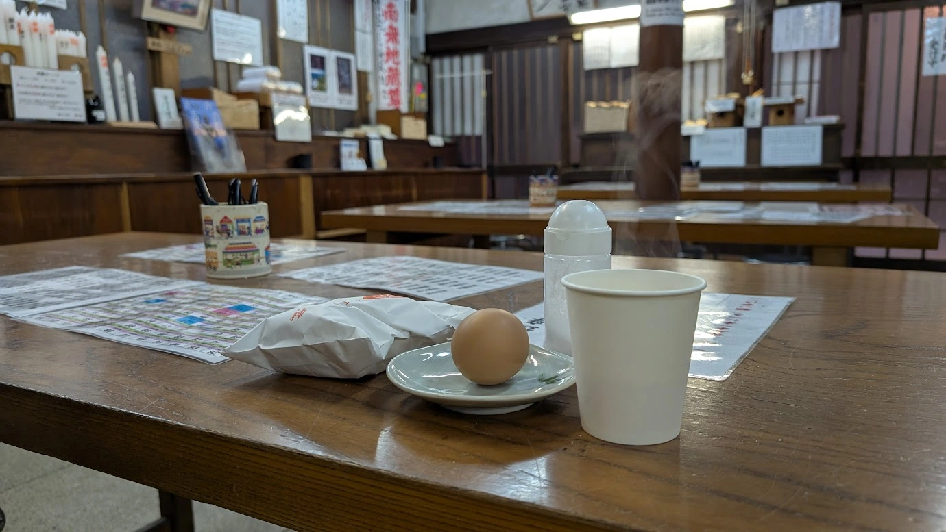 脳天大神の休憩所のテーブルに置かれた、お下がり(お供え物)のゆで卵、お餅、そして湯気が立つ温かいお茶の紙コップ。背景には祈祷受付の様子が写っている温かい雰囲気の風景。