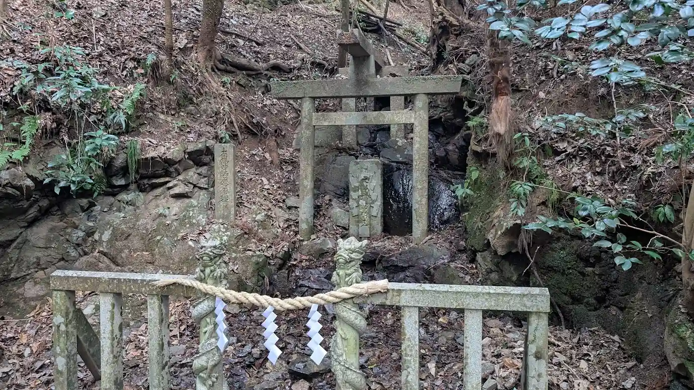 脳天大神への石段の途中にある大聖不動明王が祀られた場所。小さな石橋の奥に石鳥居と石碑があり、背後には岩肌と小さな滝(水流)が見える風景。