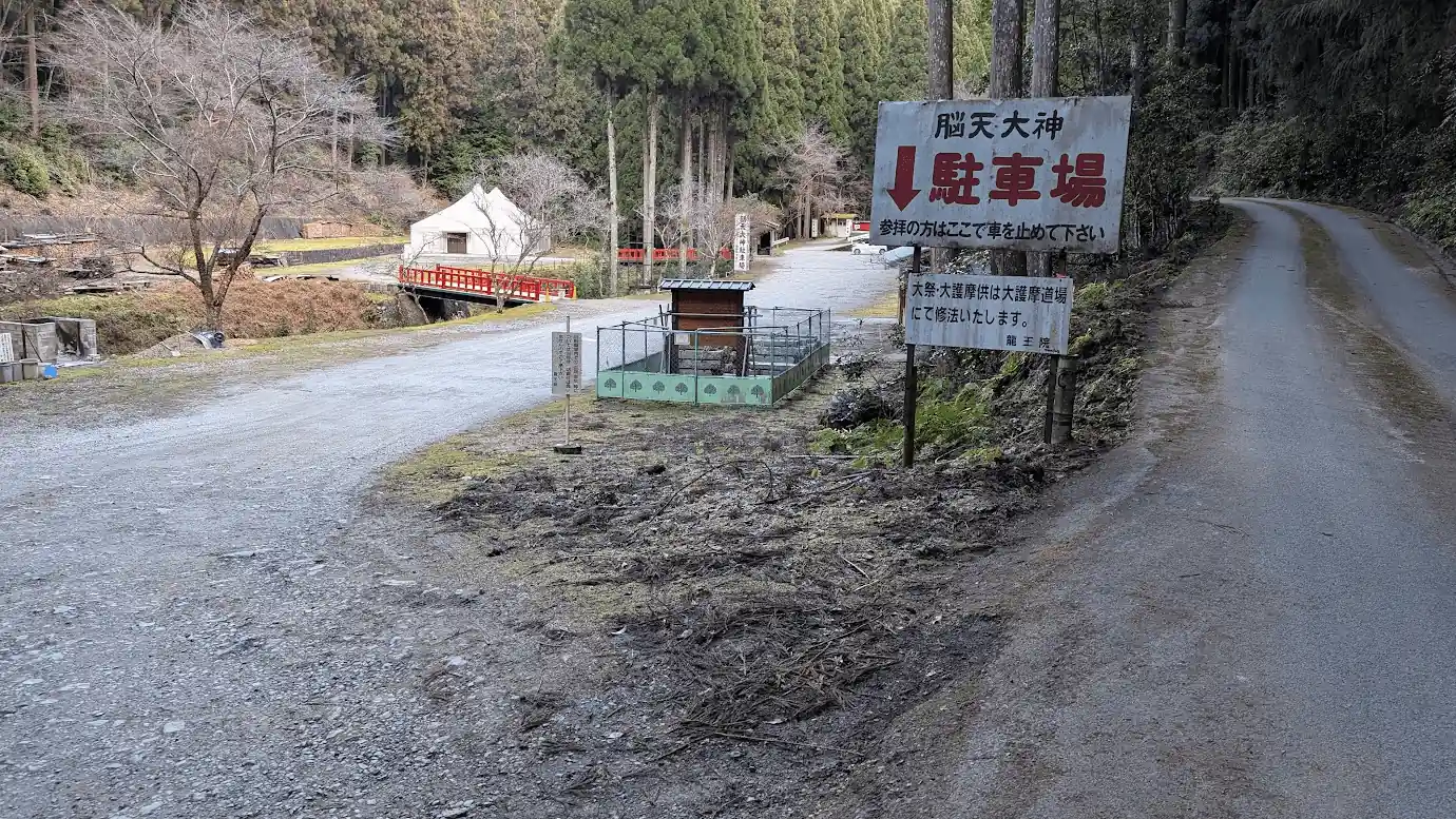 脳天大神のすぐそばにある未舗装の広い専用駐車場。「脳天大神 駐車場」と書かれた大きな看板があり、奥には境内の赤い橋が見える谷底の風景。