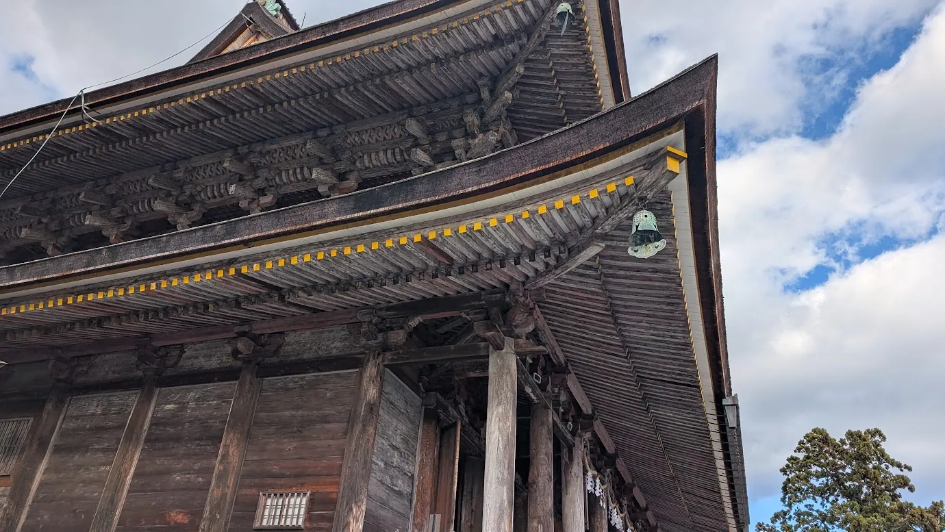 金峯山寺・蔵王堂の軒下を見上げたアップ写真。大屋根を支える複雑な木組み(組物)や規則正しく並ぶ垂木(たるき)、屋根の隅に吊るされた風鐸(ふうたく)が青空を背景に写っている。