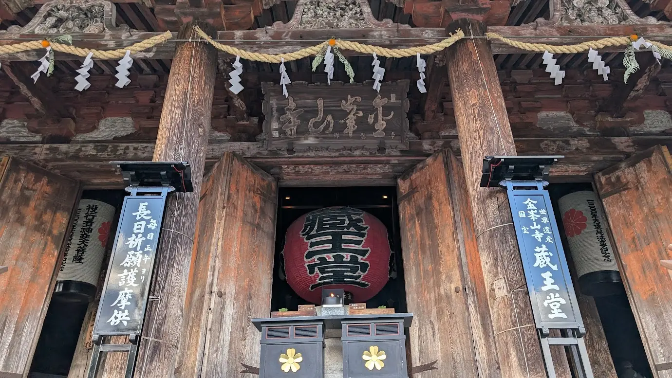 金峯山寺・蔵王堂の正面入り口のアップ写真。力強い筆致の「金峯山寺」の扁額、赤く巨大な「蔵王堂」の大提灯、注連縄、そして巨大な自然木の柱が写っている。