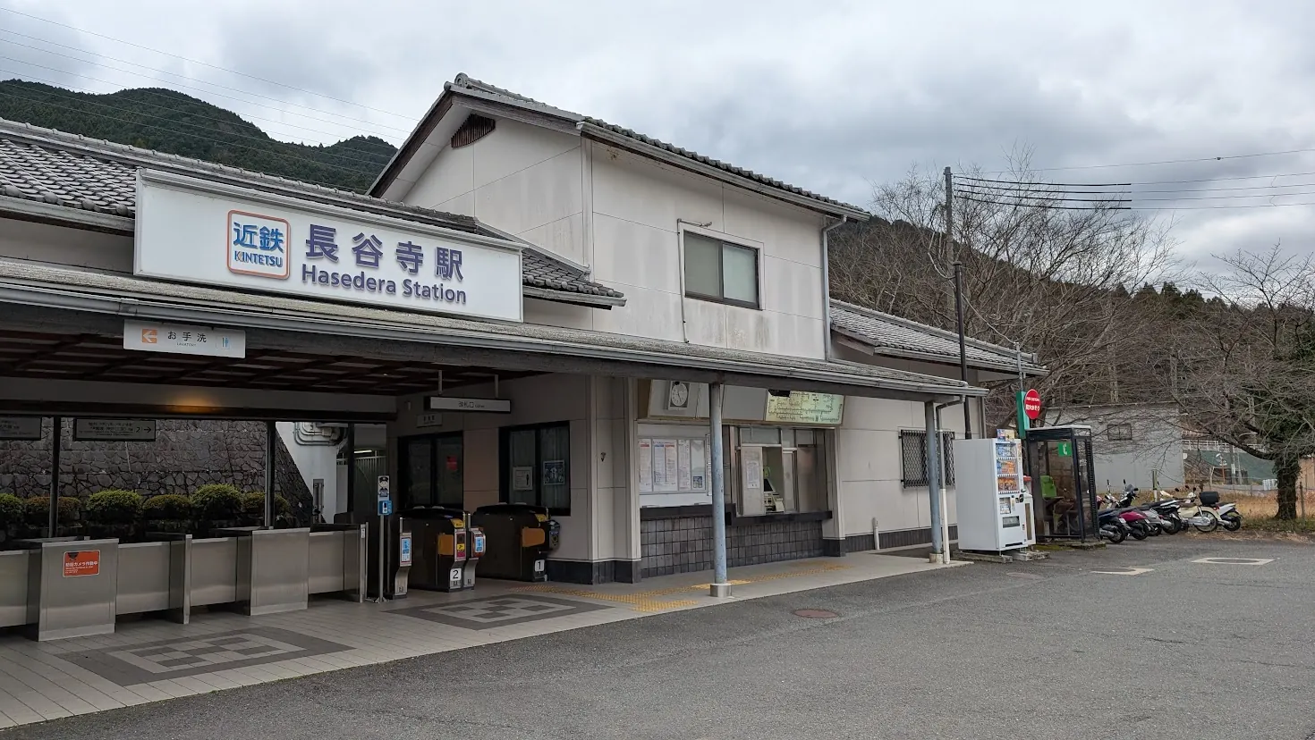 近鉄大阪線・長谷寺駅の駅舎。白い壁の平屋建てで、青い文字で「近鉄 長谷寺駅」と書かれた看板があり、背景には冬の初瀬山が広がっている。