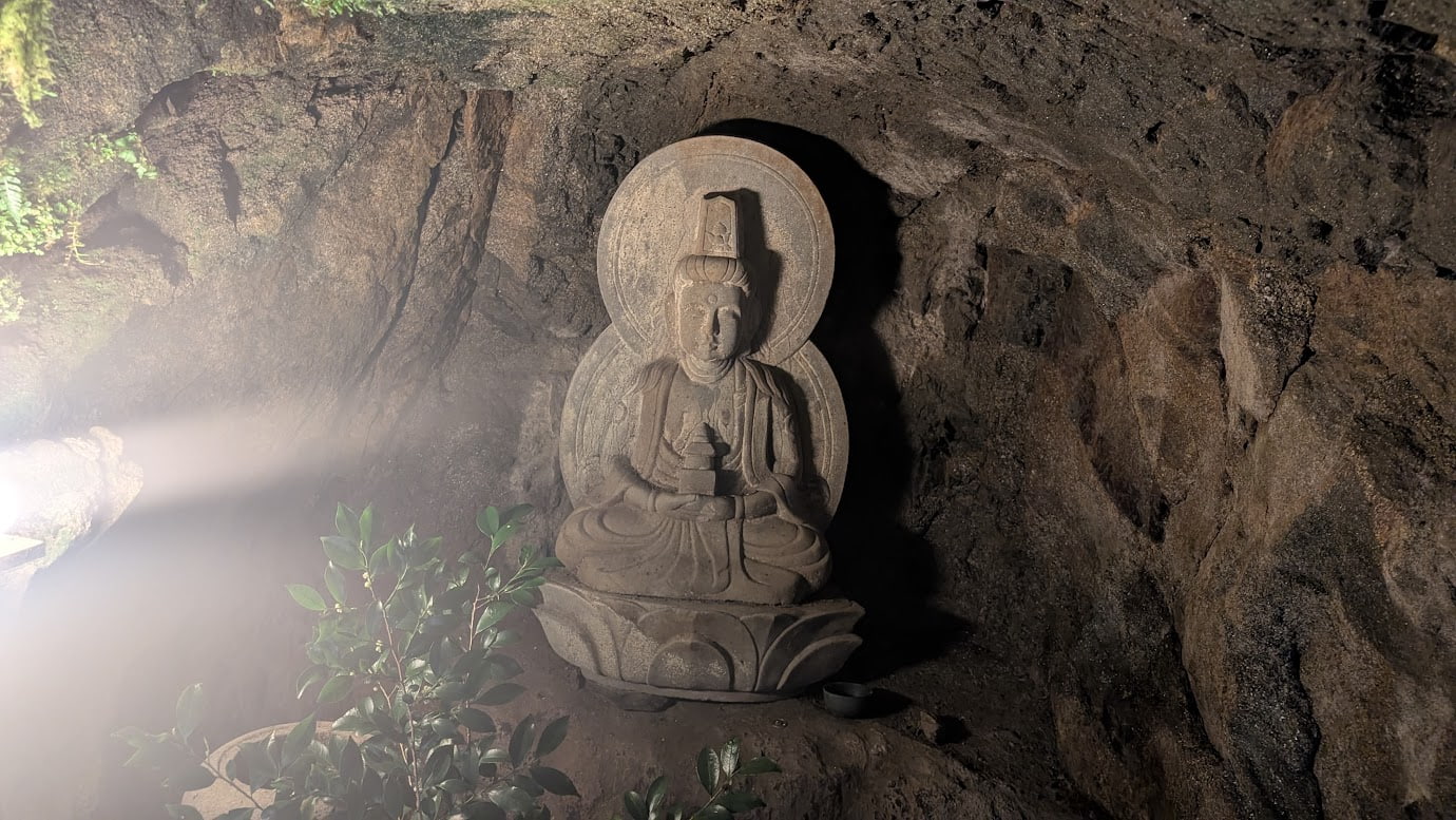 奈良県明日香村、岡寺の奥の院にある石窟(洞窟)の内部。薄暗い空間に安置された石造の弥勒菩薩坐像に、左側から神秘的な光が差し込んでいる。穏やかな微笑みを浮かべた菩薩様と、手前に供えられた緑の葉が美しい癒やしの風景(2026年3月30日撮影)。