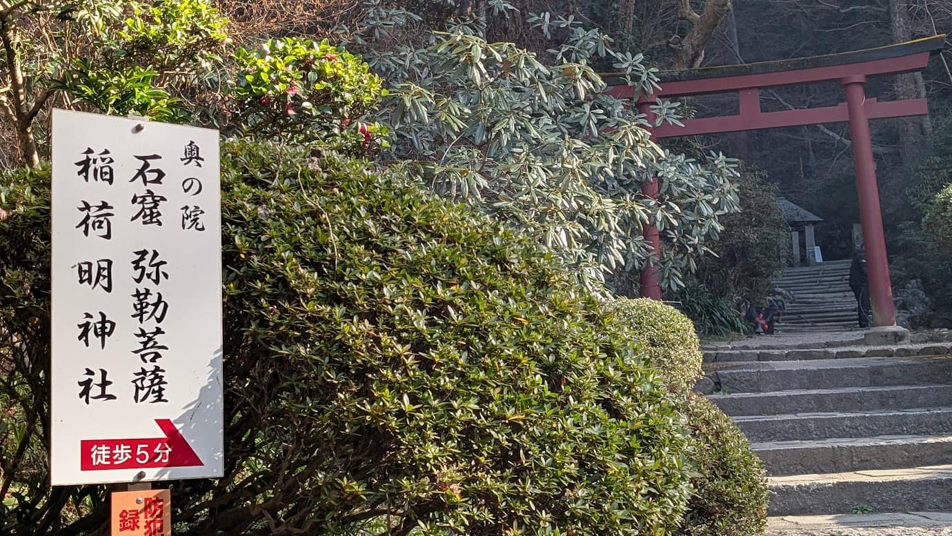 奈良県明日香村、岡寺の境内にある案内看板。「奥の院 石窟 弥勒菩薩 稲荷明神社 徒歩5分」と赤色の矢印で記されている。背景には赤い鳥居と、山へと続く石段が広がっている(2026年1月撮影)。