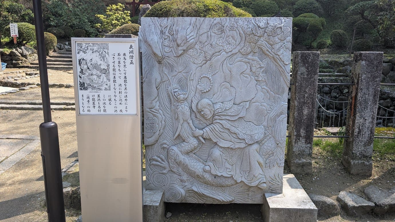 奈良県明日香村、岡寺にある「龍蓋池」の前の石碑。義淵僧正が悪龍を池に封じ込める様子が彫られた精緻な石のレリーフ。左側には、寺名の由来となった伝説を解説する白い案内板が立っている(2026年3月30日撮影)。