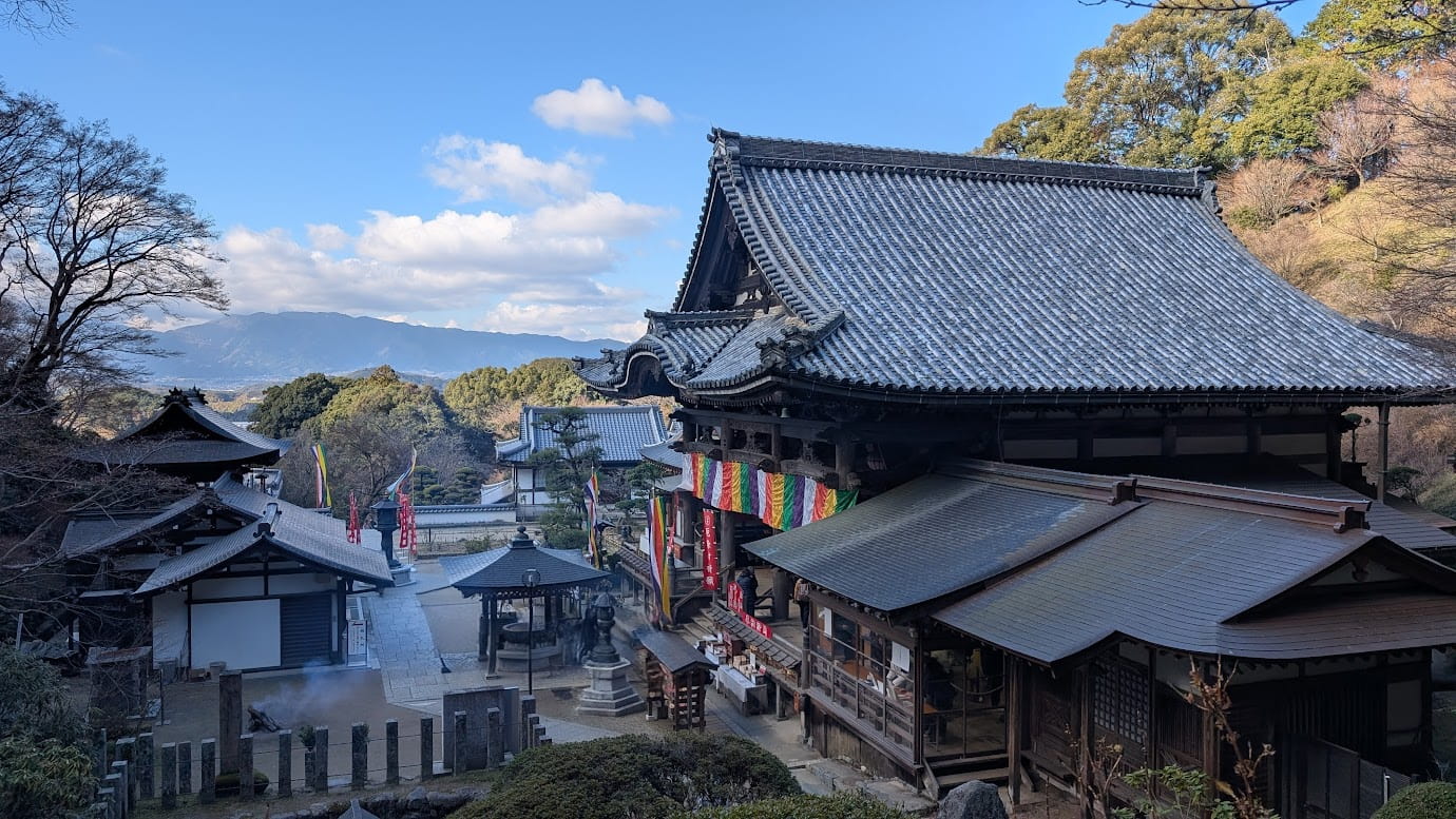 奈良県明日香村、岡寺の境内にある石畳の歩道。手前には本堂の大きな屋根があり、奥には飛鳥の山々と青い空が広がっている。新春の光に包まれた、静かで開放感のある境内の風景(2026年1月撮影)。