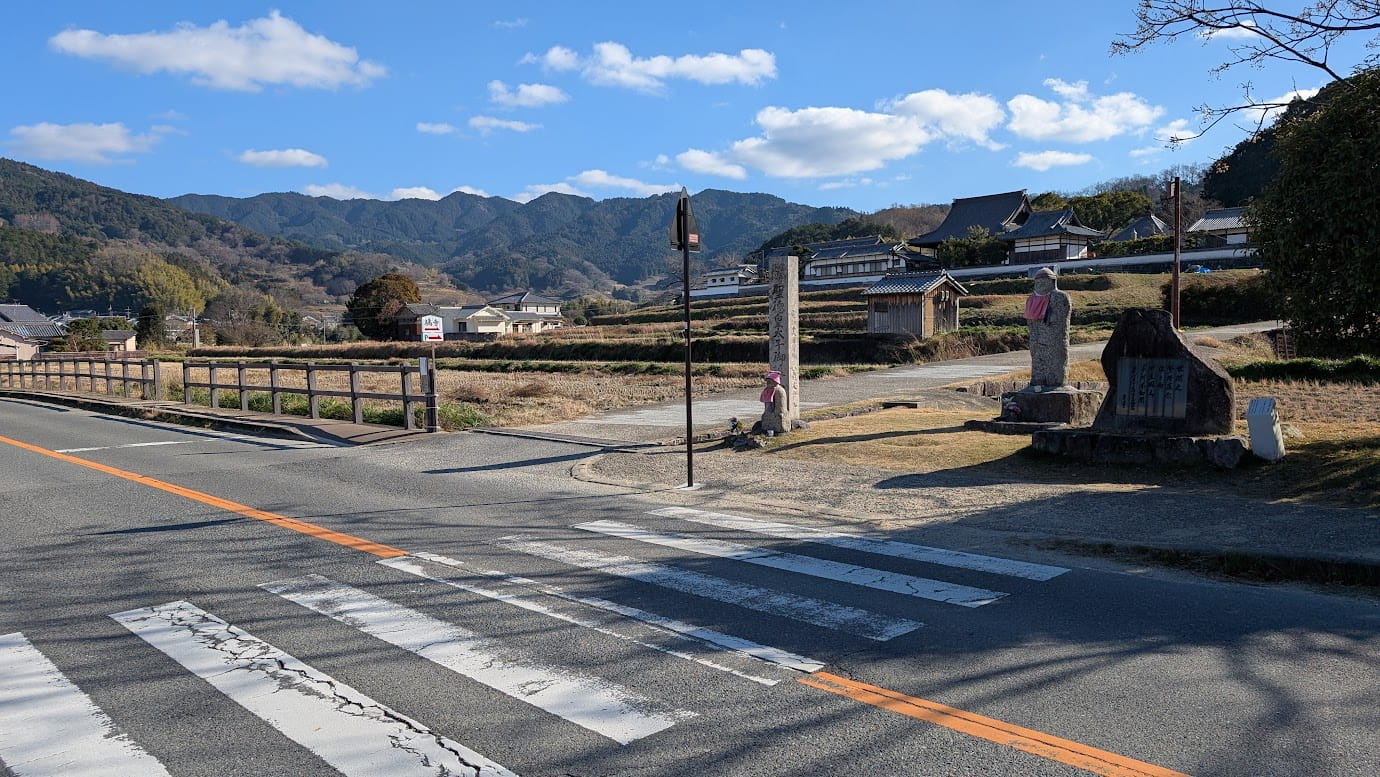 奈良県明日香村、橿原方面から橘寺へ続く道路。前方に横断歩道と、左手に橘寺の入り口を示す案内看板や石碑が見える分岐点の風景（2026年1月12日撮影）。