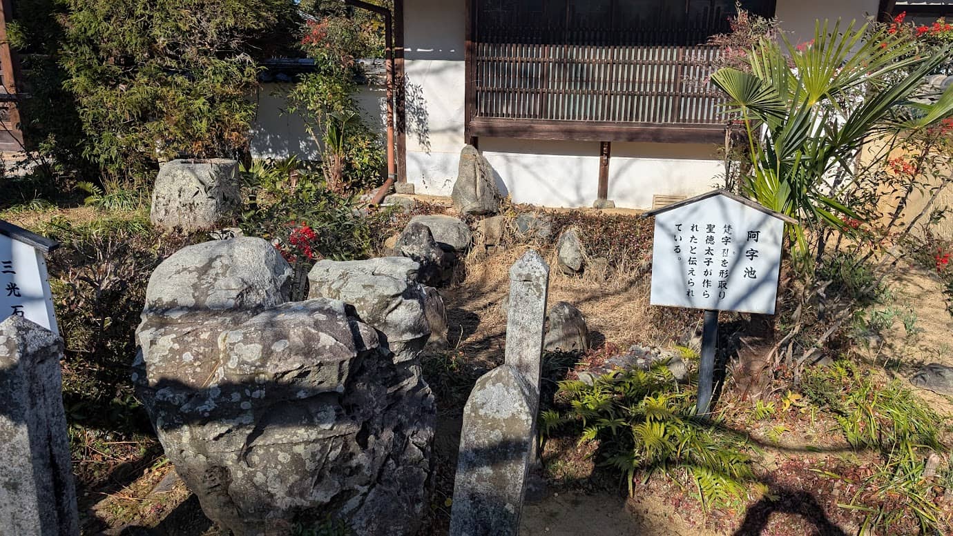 奈良県明日香村・橘寺の境内にある「阿字池」と「三光石」。梵字の「阿」の形をした古池と、その周囲に配された大きな石、そして解説板が冬の光に照らされている風景。