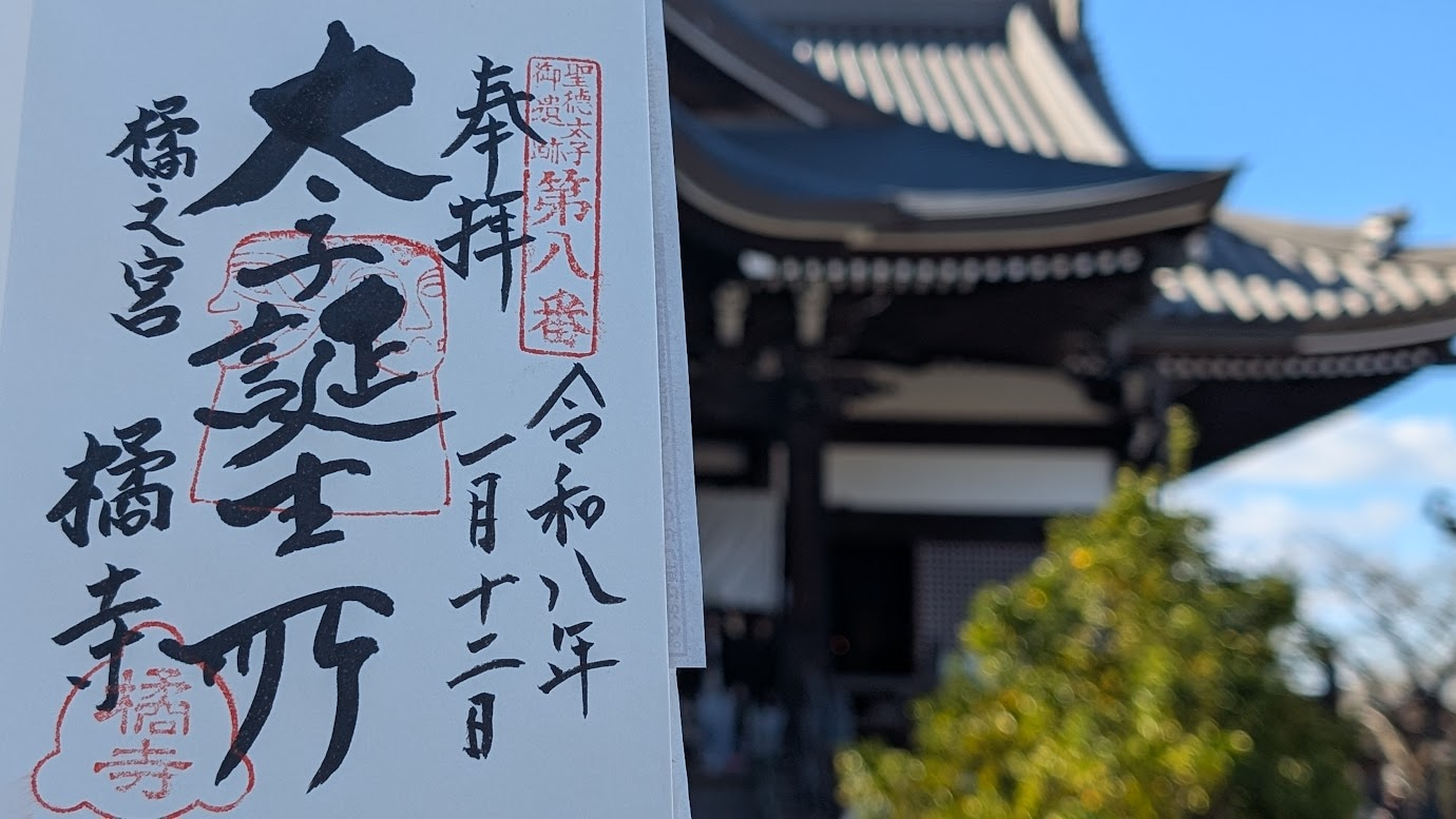 奈良県明日香村の橘寺。手前に「聖徳太子 誕生所」と墨書きされた御朱印帳を掲げ、背景には聖徳太子を祀る本堂（太子殿）が静かに佇んでいる（2026年1月12日拝受）。