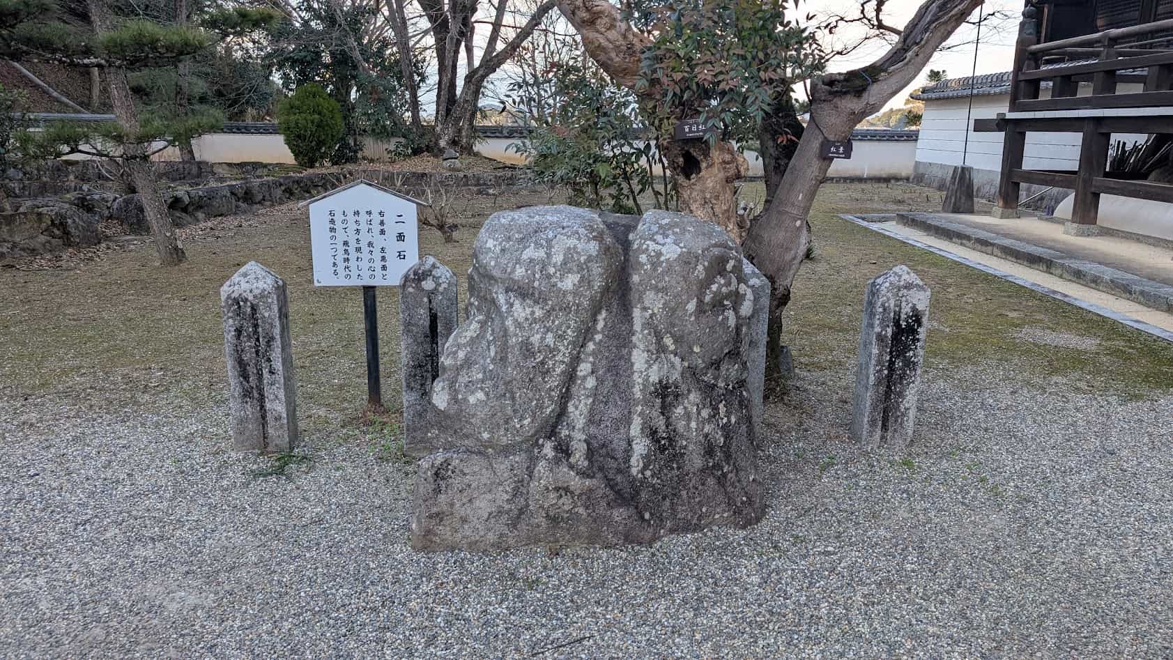 奈良県明日香村の橘寺境内にある「二面石」。一つの大きな石の両面に、善と悪の二つの顔が彫られた飛鳥時代の石造物。背景には解説板と境内の松の木が見える。