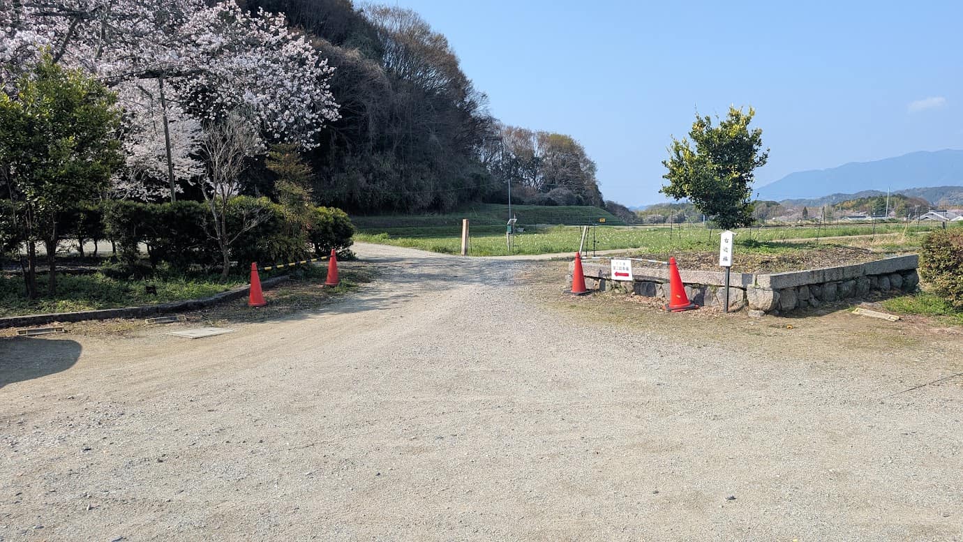奈良県明日香村、橘寺の第一駐車場奥にある第二駐車場への入口。赤いカラーコーンと「すぐ奥 第二駐車場」と書かれた赤い矢印の案内板が設置されており、砂利道の先に広がる駐車スペースへと誘導している風景（2026年3月30日撮影）。
