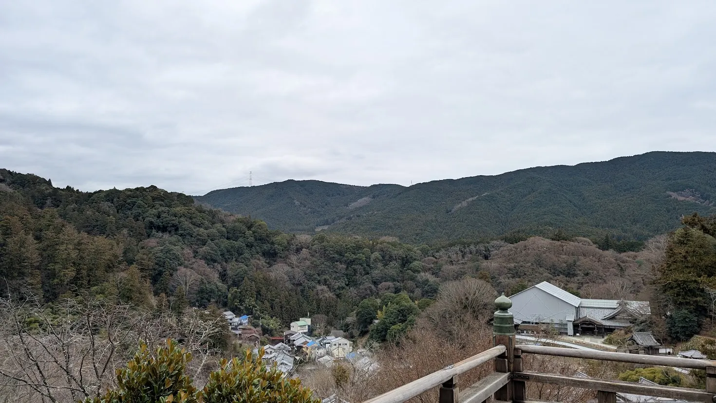 長谷寺の本堂（舞台）の欄干越しに広がる冬の風景。手前には門前町の家々、奥には初瀬の深い山々が連なり、どんよりとした冬空の下で静寂に包まれている。