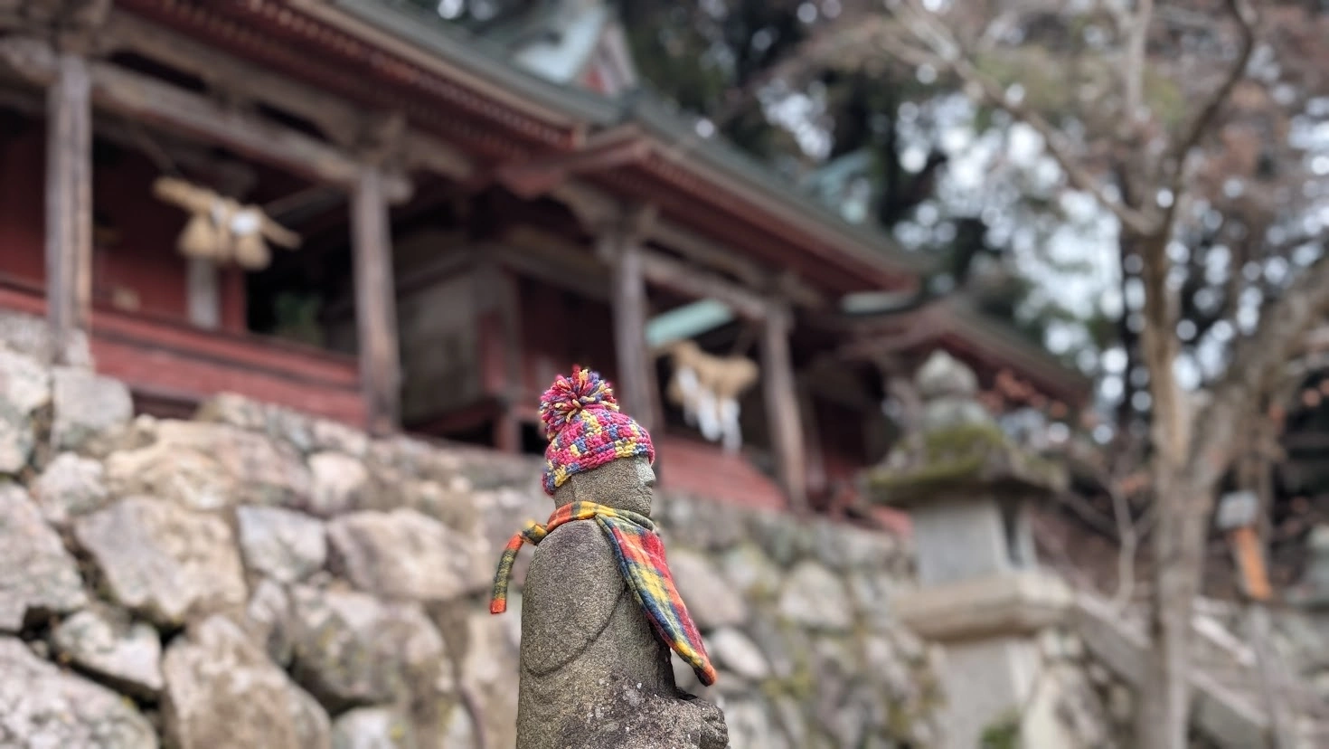 長谷寺の境内に佇む、手編みの赤いニット帽と前掛けを身につけた小さなお地蔵様。背景には冬の柔らかな光に包まれた木造の摂社がボケて写っている。
