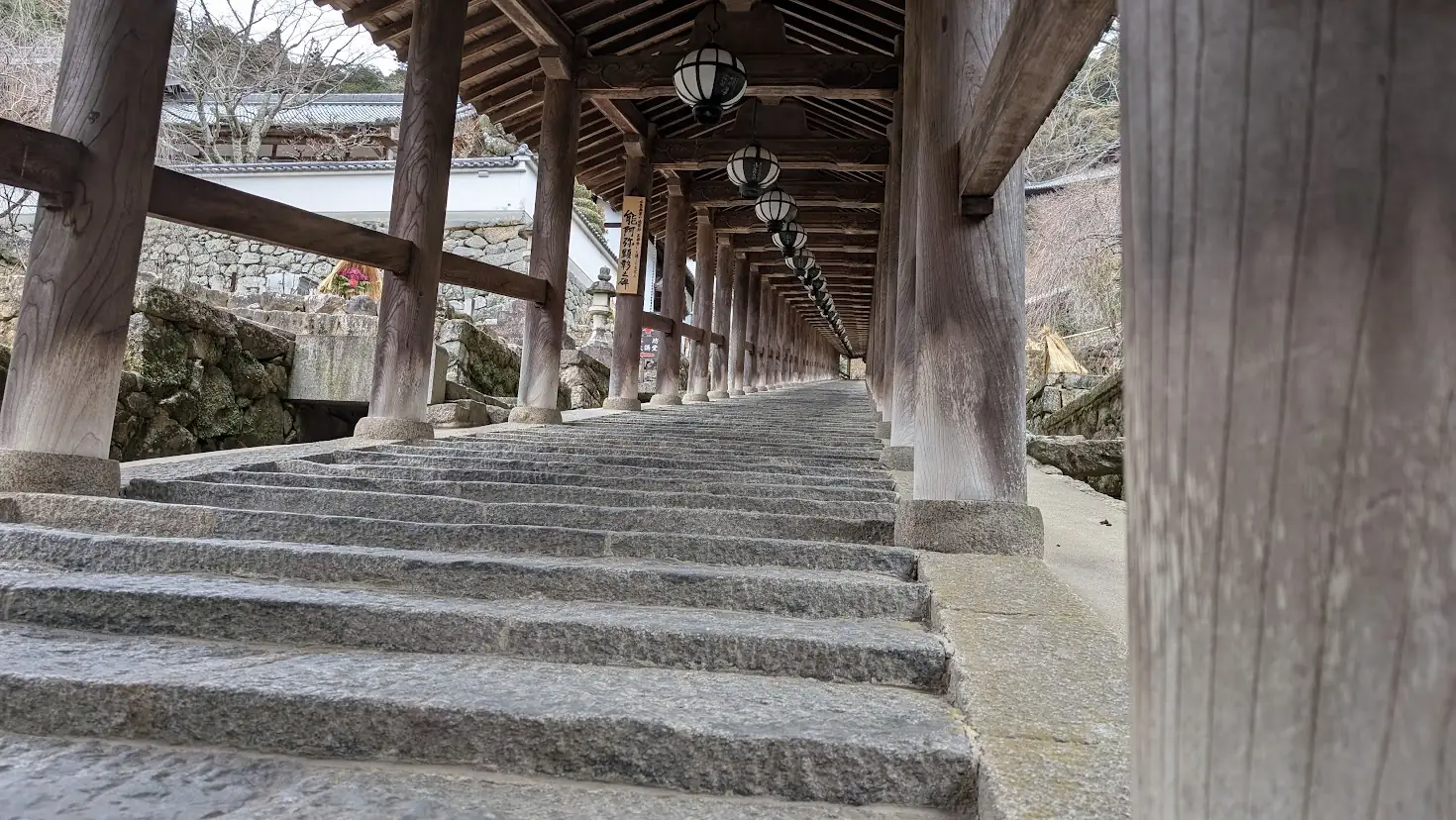 長谷寺の登廊を低い位置から撮影した写真。手前に石段の質感が大きく写り、屋根を支える太い木柱と天井の灯笼が、上方の本堂へと向かって美しく連なっている。