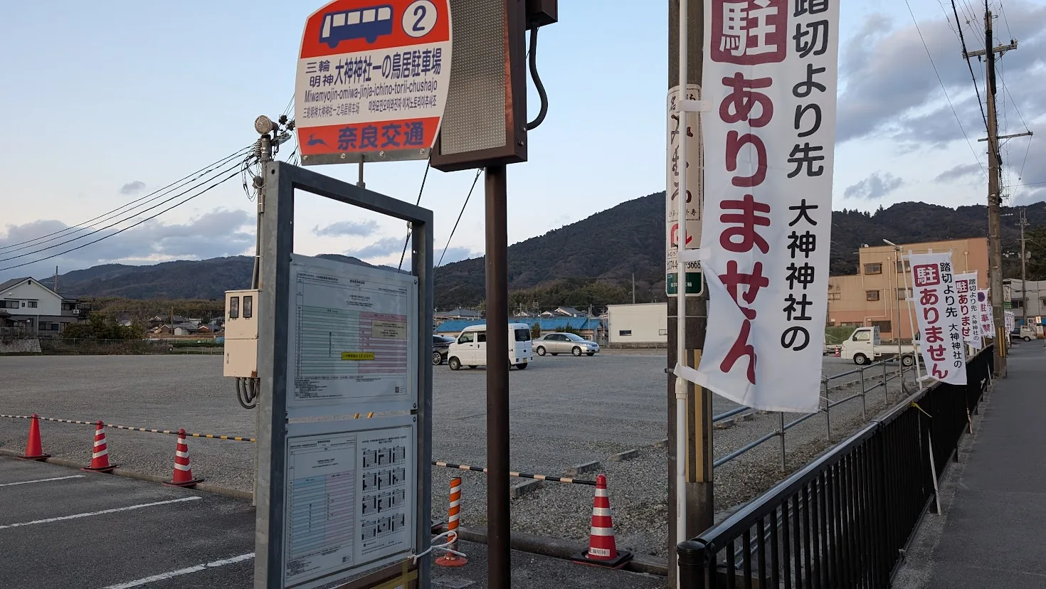 夕暮れ時、三輪山を背景にした大神神社一の鳥居駐車場のバス停(奈良交通)。周囲には「踏切より先駐ありません」の幟が立つ風景。