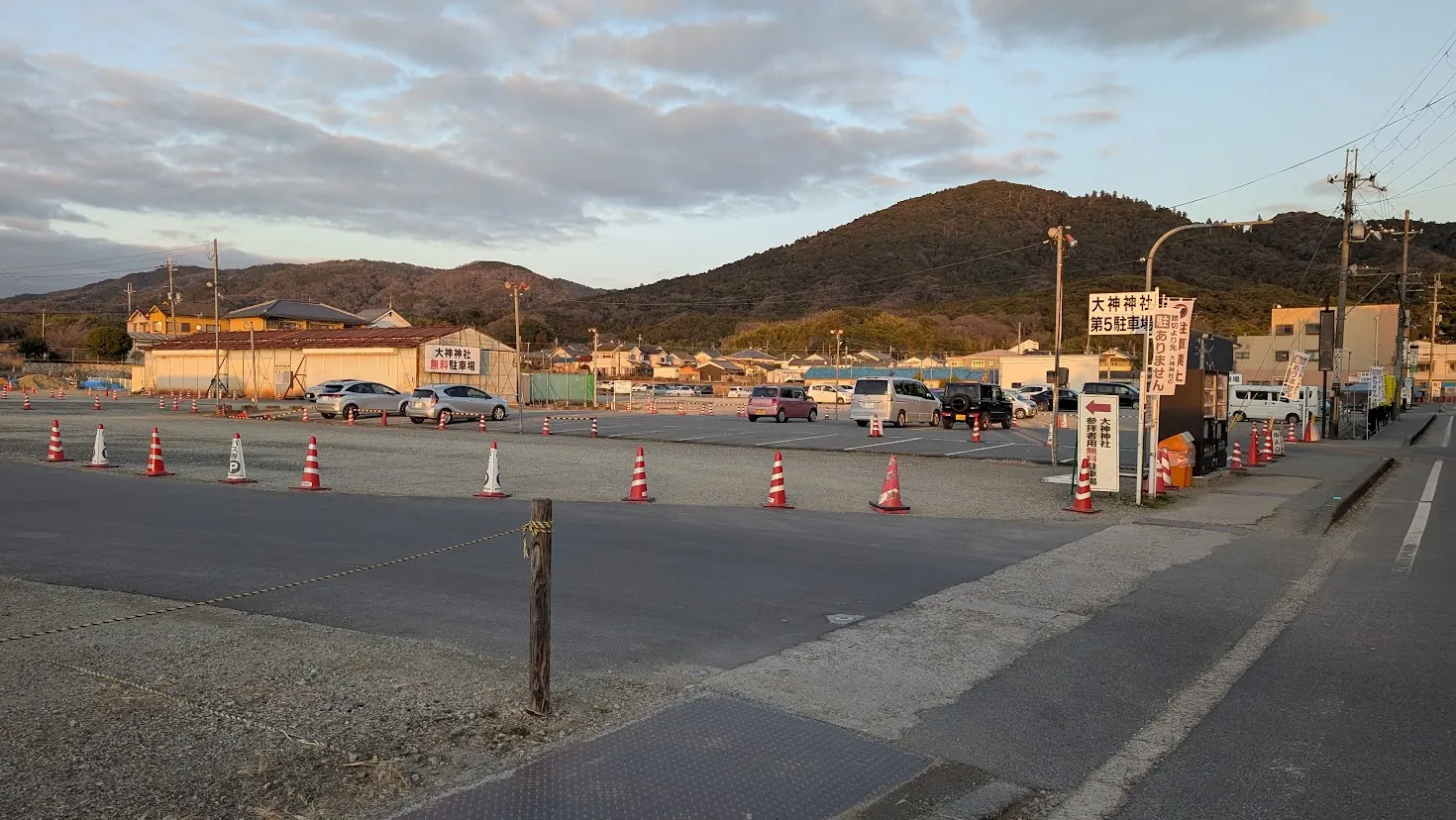 夕暮れ時、三輪山を背にした大神神社の広大な第五駐車場。舗装され、赤いコーンで区切られた駐車スペースの風景。