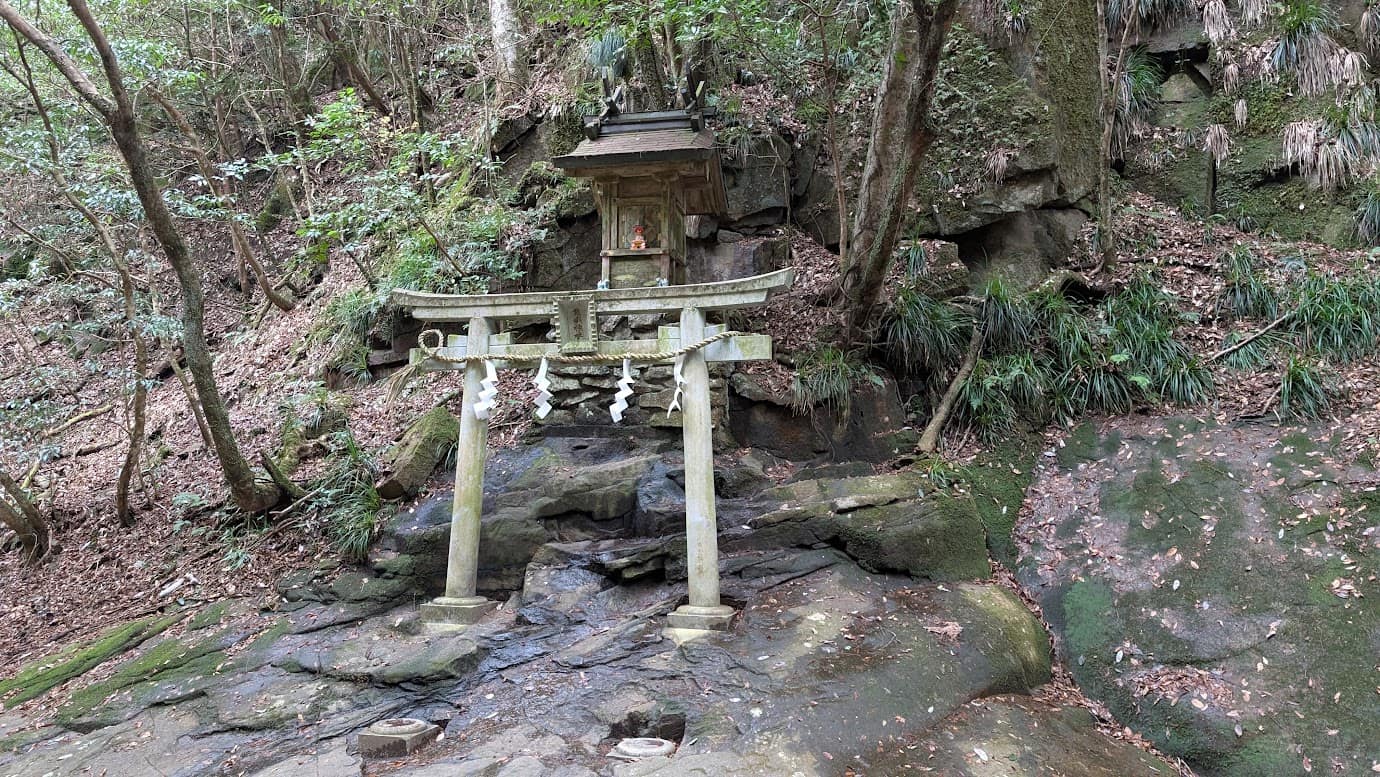 奈良県宇陀市の龍鎮神社。大きな岩肌の上に立つ小さな石造りの鳥居と、その背後に安置された木造の小さな祠（拝殿）。周囲は深い森に囲まれ、厳かな雰囲気が漂っている。