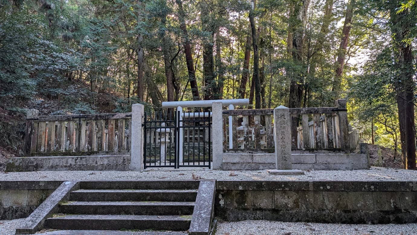 奈良県吉野町、山に囲まれた静かな場所にある後醍醐天皇の御陵(塔尾陵)。石造りの低い柵に囲まれた聖域の中に、白い鳥居と石灯籠が見える。手前には石段があり、右側には「後醍醐天皇 塔尾陵」と刻まれた石柱が立っている(2026年1月撮影)。