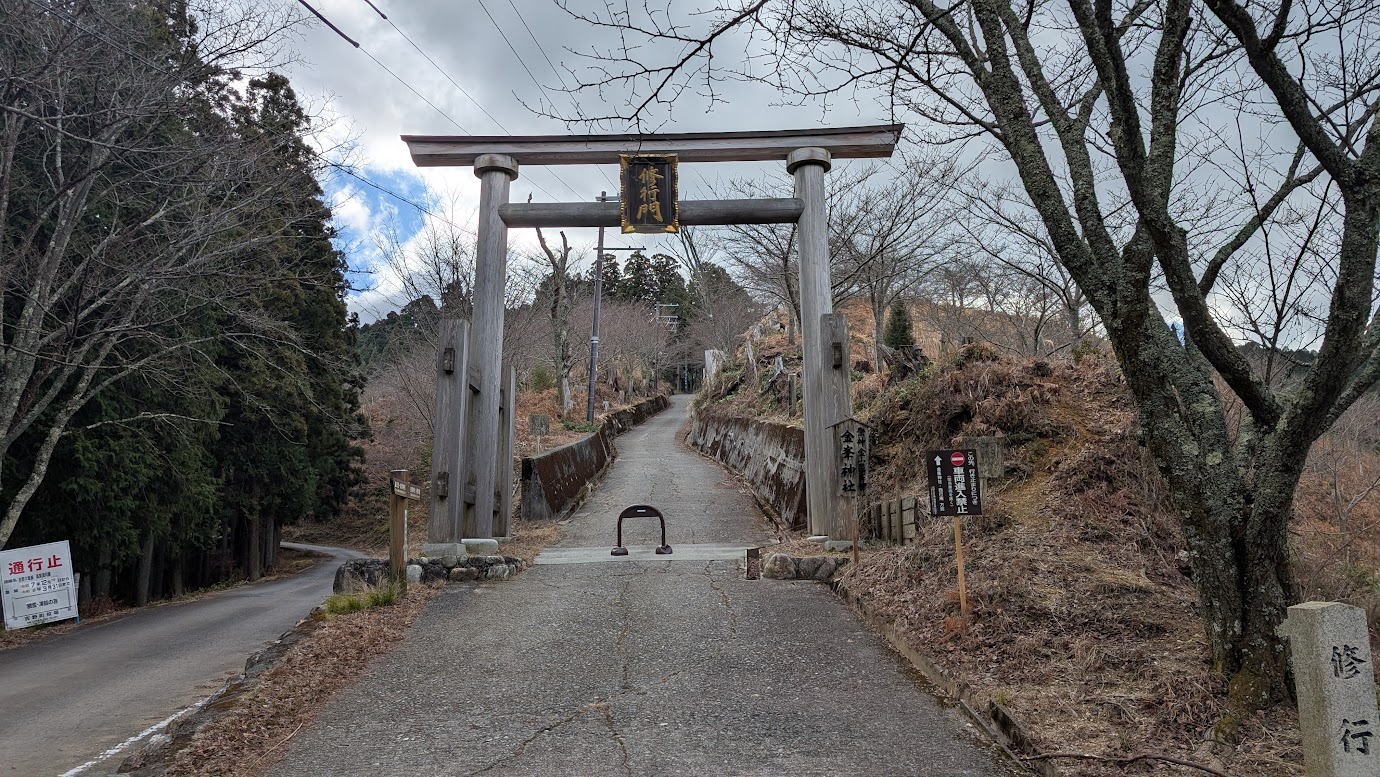 奈良県吉野町、吉野山の最深部・奥千本にある金峯神社の修行門(木造鳥居)。鳥居の扁額には「修行門」とあり、その先には車両進入禁止の急な上り坂が続いている。冬の枯れ木に囲まれた、峻烈な空気感漂う風景(2026年1月撮影)。