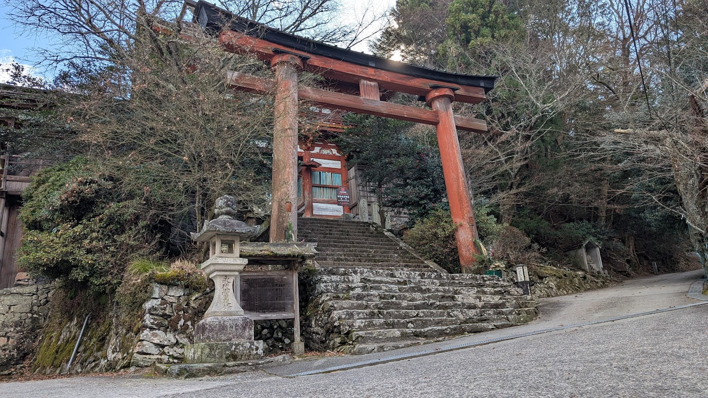 奈良県吉野町、世界遺産・吉野水分神社の入り口に立つ巨大な朱色の木造鳥居。急な坂道の石段の上に建ち、周囲を冬の木々に囲まれている。鳥居の奥には楼門が見える静謐な風景（2026年1月撮影）。