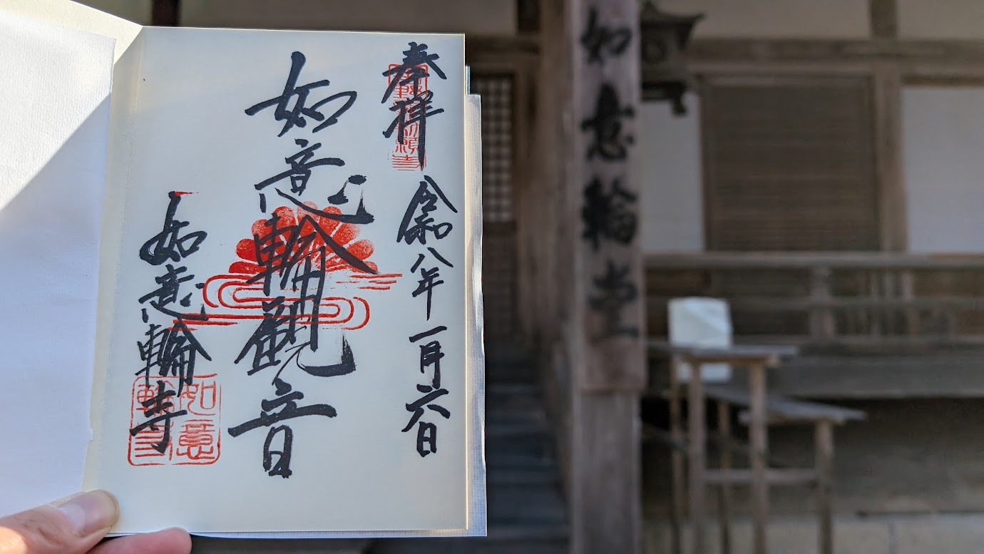奈良県吉野町、如意輪寺の御朱印。中央に「如意輪観音」と墨書きされ、菊の御紋の朱印が押されている。背景には如意輪寺の本堂（如意輪堂）がぼかして写っている（2026年1月6日拝受）。