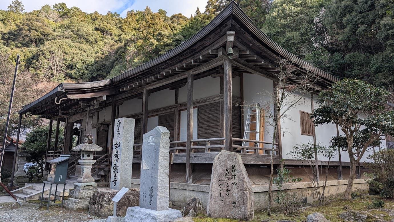 奈良県吉野町、如意輪寺の本堂。手前には「浄土宗 如意輪寺」と刻まれた寺標と、松尾芭蕉の句碑が立っている。入母屋造の落ち着いた本堂が、背後の深い山林を背に佇む新春の風景（2026年1月撮影）。