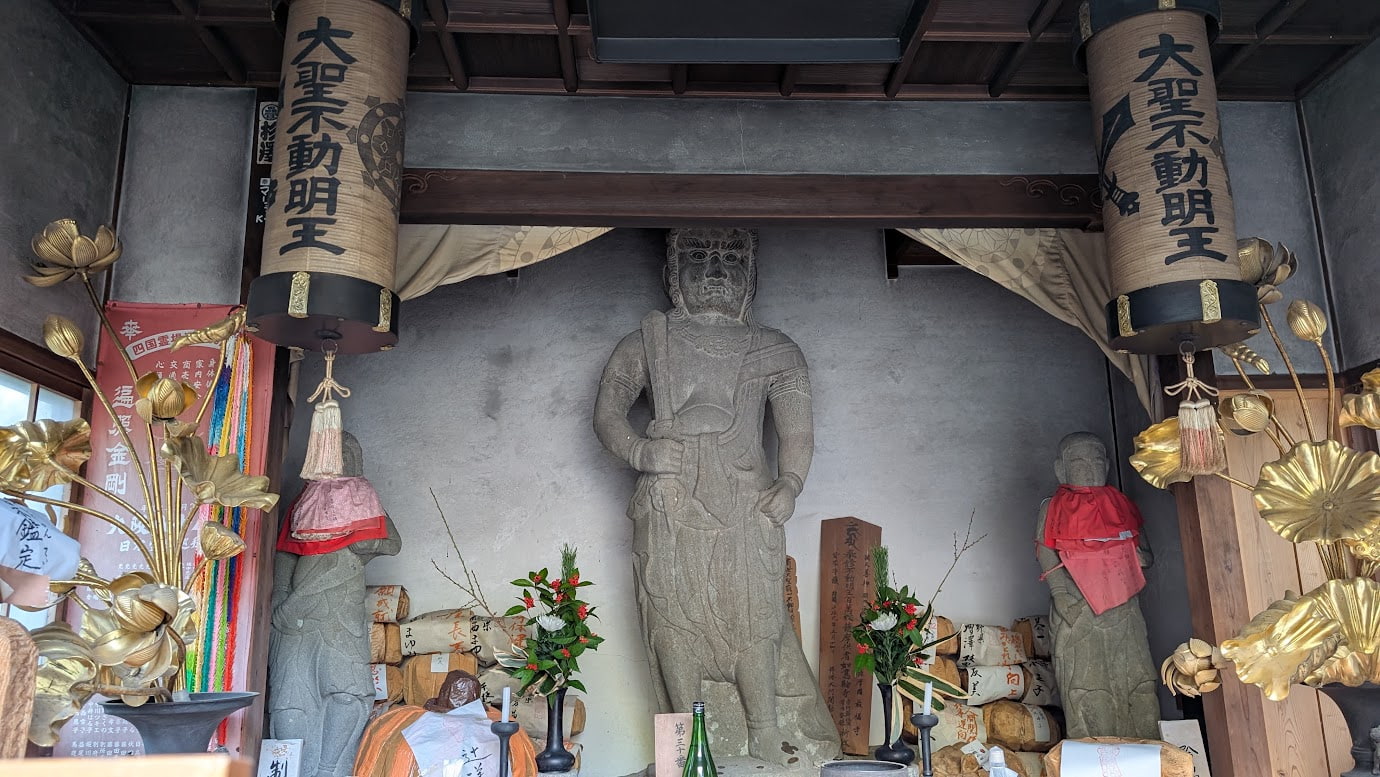 奈良県吉野町、如意輪寺の境内にある難切不動尊。中央に石造の不動明王立像が立ち、左右には赤い布を掛けられた脇侍(矜羯羅童子・制咤迦童子)が控えている。上部には「大聖不動明王」と書かれた大きな提灯が吊るされている風景(2026年1月撮影)。