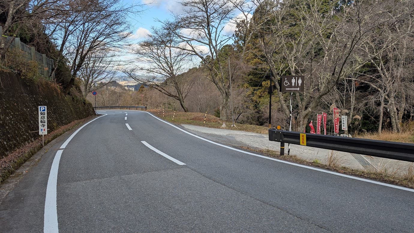 奈良県吉野町、如意輪寺の無料駐車場入口。下り坂の途中にあり、右側には「如意輪寺 駐車場入口」と書かれた看板と、浄土宗の旗、歴史的な説明看板が立っている。冬の静かな山道の風景（2026年1月撮影）。