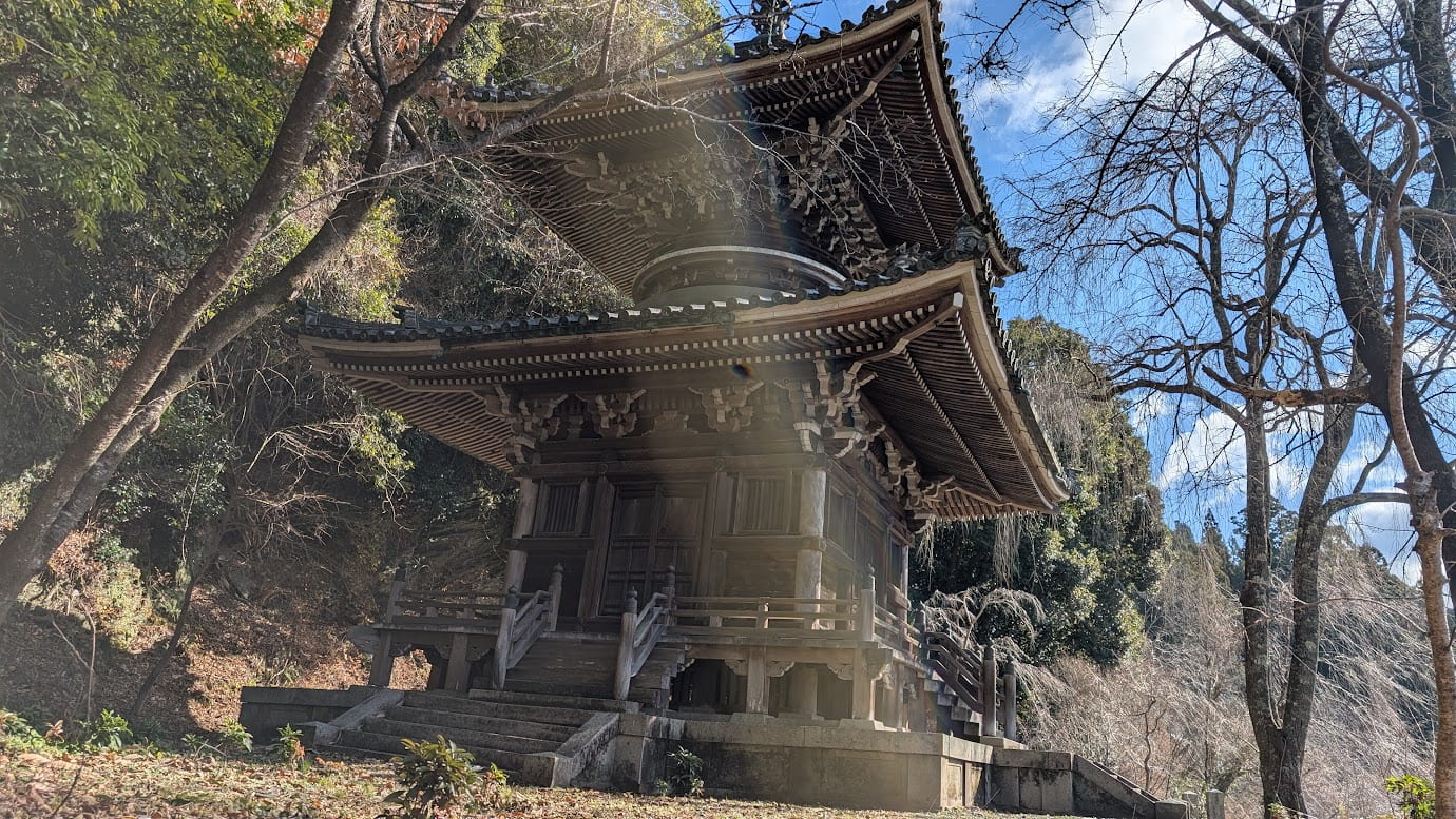 奈良県吉野町、如意輪寺の多宝塔。二層の木造建築で、上層が円形、下層が方形の特異な構造。周囲を冬の木々に囲まれ、太陽の光がレンズフレアとなって差し込む静謐な風景（2026年1月撮影）。