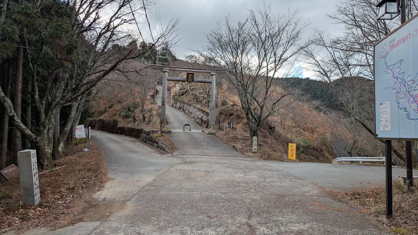 山道の終点。前方に「修行門」と書かれた木造の鳥居が見えてくる。左側には舗装路が続き、右側には観光案内マップの看板が立っている風景(2026年1月撮影)。