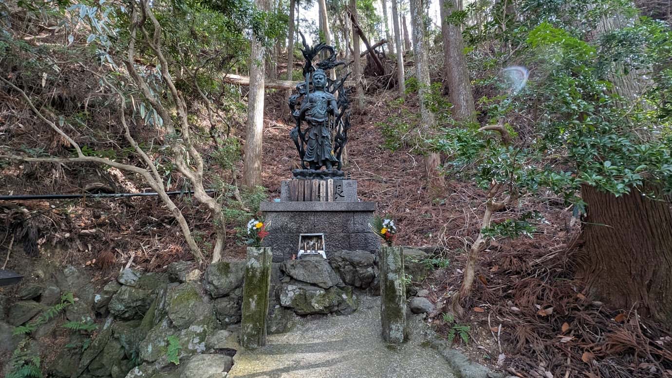 吉野山の山道脇、斜面の切り立った岩場に安置された黒い不動明王像。背後には炎を背負い、力強い剣を携えて参拝者を見守っている(2026年1月撮影)。