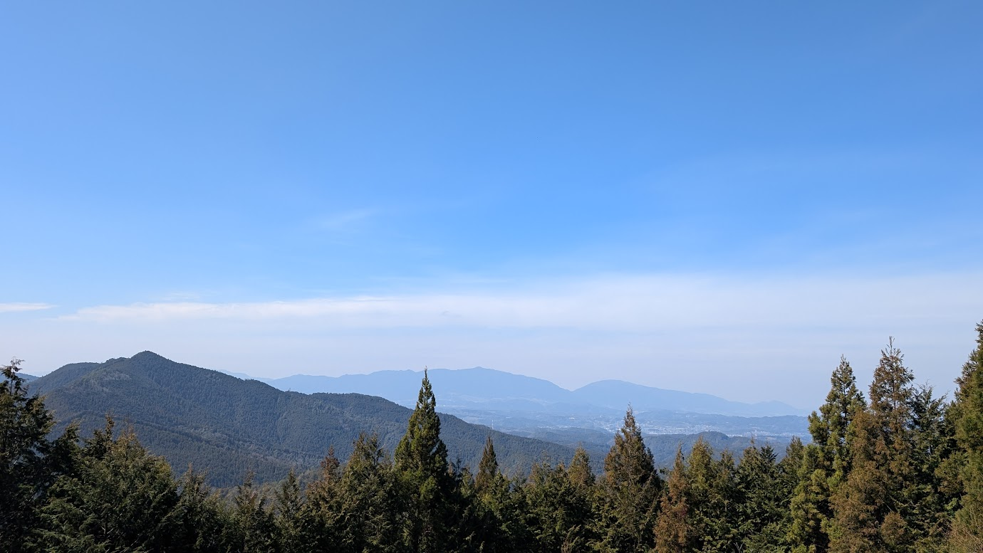 鳳閣寺へ向かう山道から見下ろす奈良県吉野地方の景色。幾重にも重なる青い山並みと、遠くに広がる街並み、澄み渡った青空が広がっている。手前には深い杉林の頂部が見える。