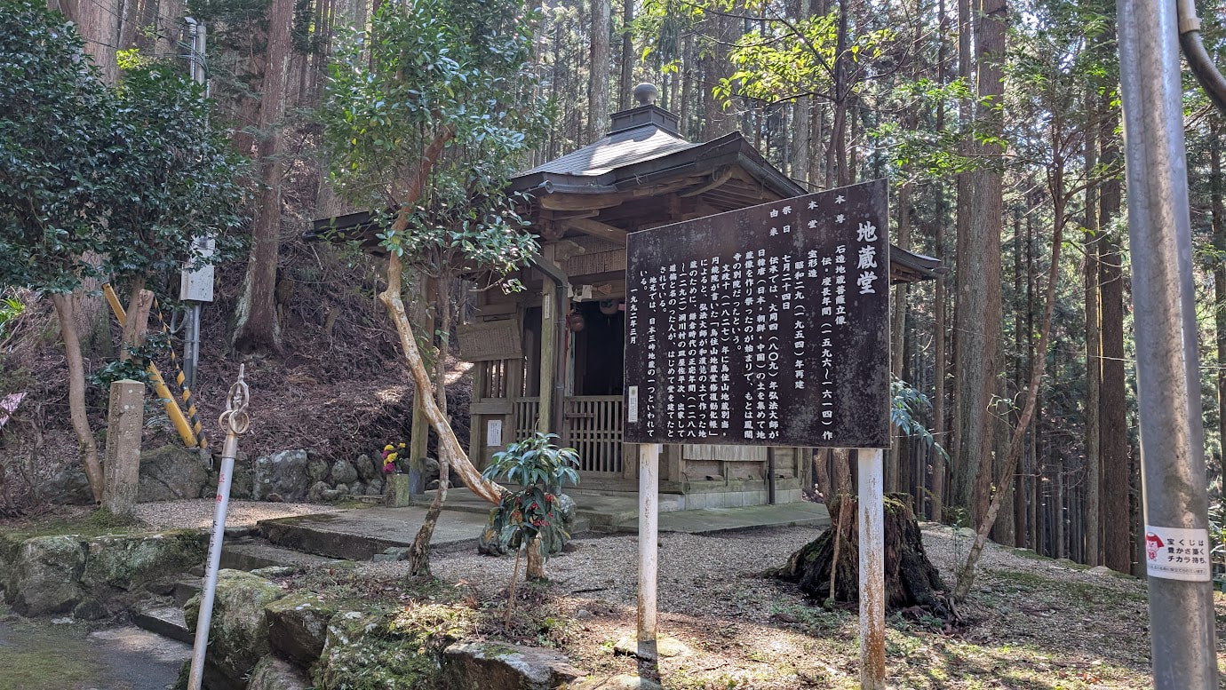 奈良県黒滝村、鳳閣寺の下にある地蔵堂の全景。手前には大きな黒い案内板があり、奥に木造の小さなお堂が建っている。周囲は高い杉の木に囲まれている。