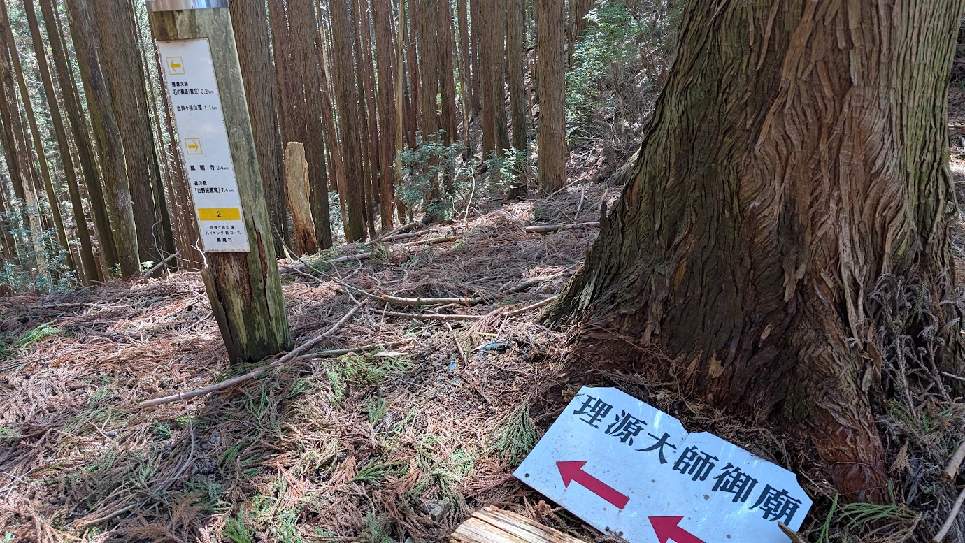 鳳閣寺の裏山、廟塔へと続く険しい山道の途中に立つ案内板。地面には「理源大師御廟」と赤い矢印が書かれた白い看板があり、横の木の柱には百貝岳や鳳閣寺までの距離が記されている。