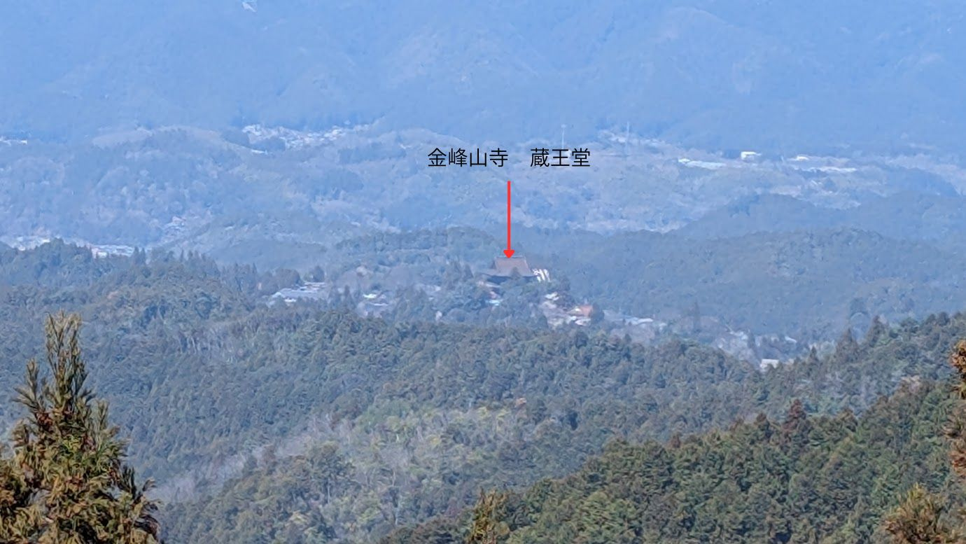 鳳閣寺の展望台から遠くに見える金峯山寺 蔵王堂のアップ画像。山々の連なりの向こう側に、蔵王堂の大きな檜皮葺の屋根がはっきりと見えている。赤い矢印で場所が示されている。
