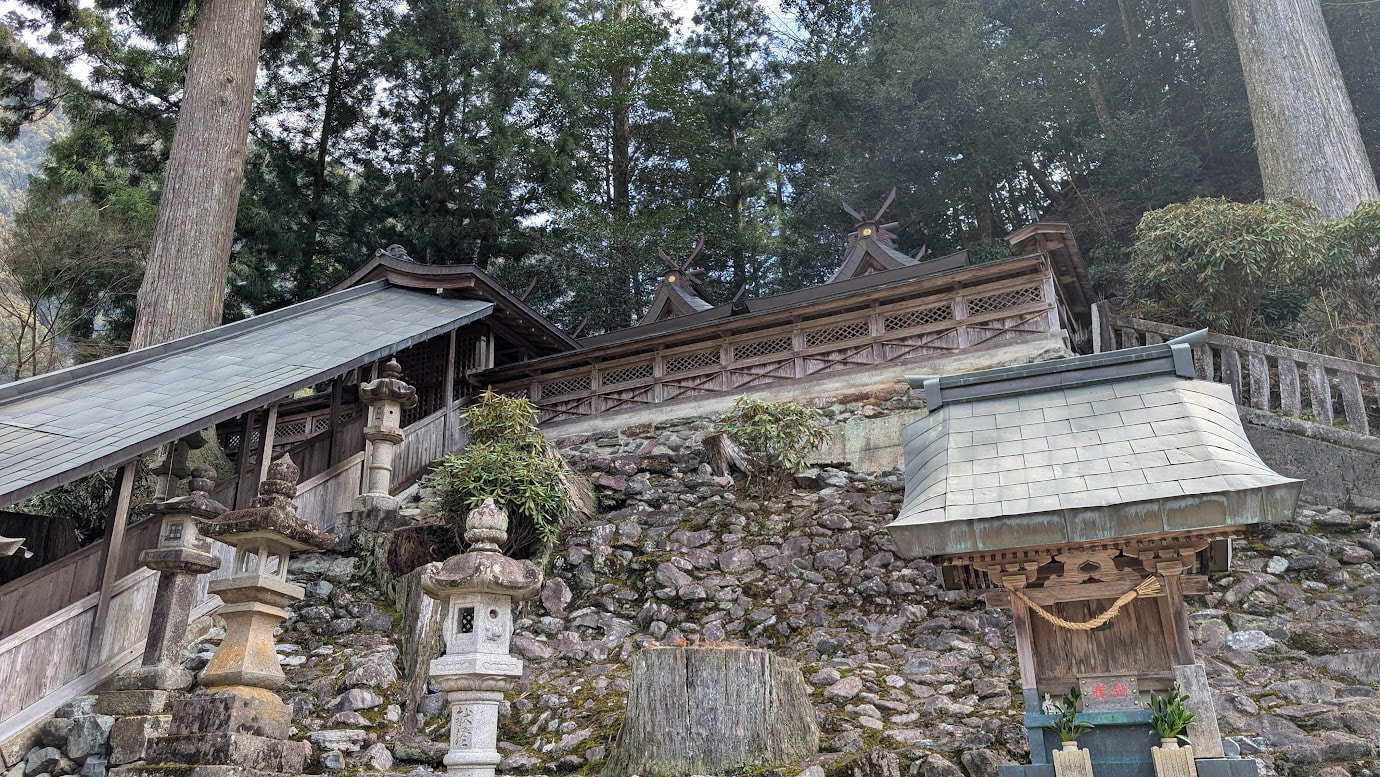 奈良県黒滝村の河分神社。苔むした境内に、素朴な木造の摂社が立っている。背後には、本殿の屋根が少しだけ見え、さらに奥には深い森が広がっている。木漏れ日が地面の苔を照らしている。