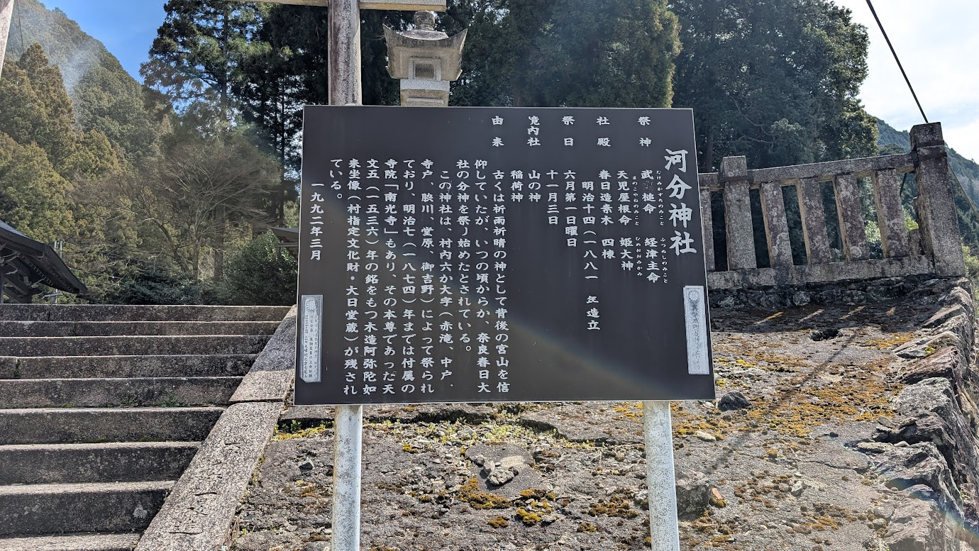 奈良県黒滝村の河分神社にある解説板。御祭神(武甕槌命など)、由緒、祭日などが詳しく記されている。背景には神社の石段と石垣が見える。