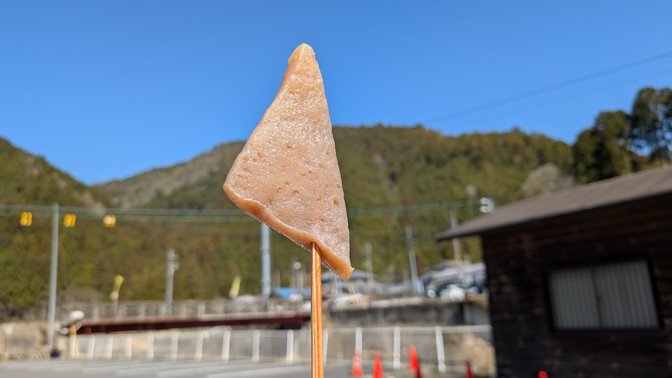 奈良県黒滝村の道の駅で販売されている名物の串こんにゃく。三角形のこんにゃくが串に刺さっており、背景には青空と山々、道の駅の建物が広がっている。