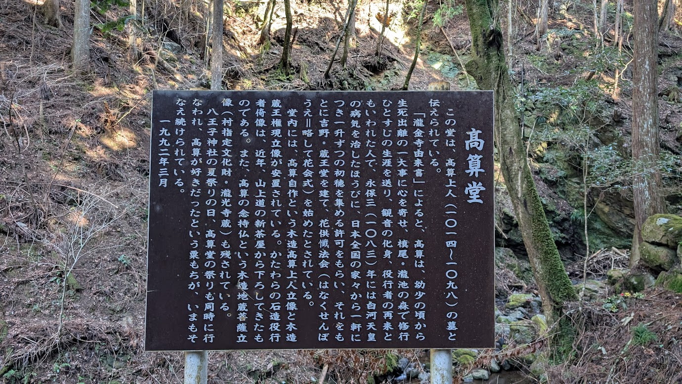 瀧光寺にある高算堂の解説板。高算上人の生涯や白河天皇との関わり、本尊の石造役行者像などについて、黒地に白い文字で詳しく書かれている。背景には苔むした石垣と森が見える。