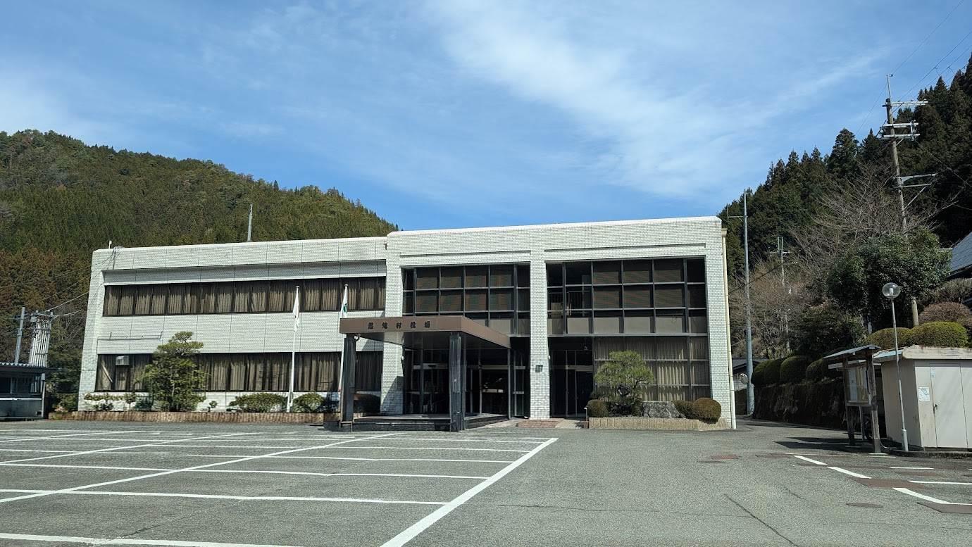 奈良県黒滝村役場の外観。白いタイルの2階建て庁舎で、手前には広い駐車場がある。背景には奥吉野の深い緑の山々と澄んだ青空が広がっている。