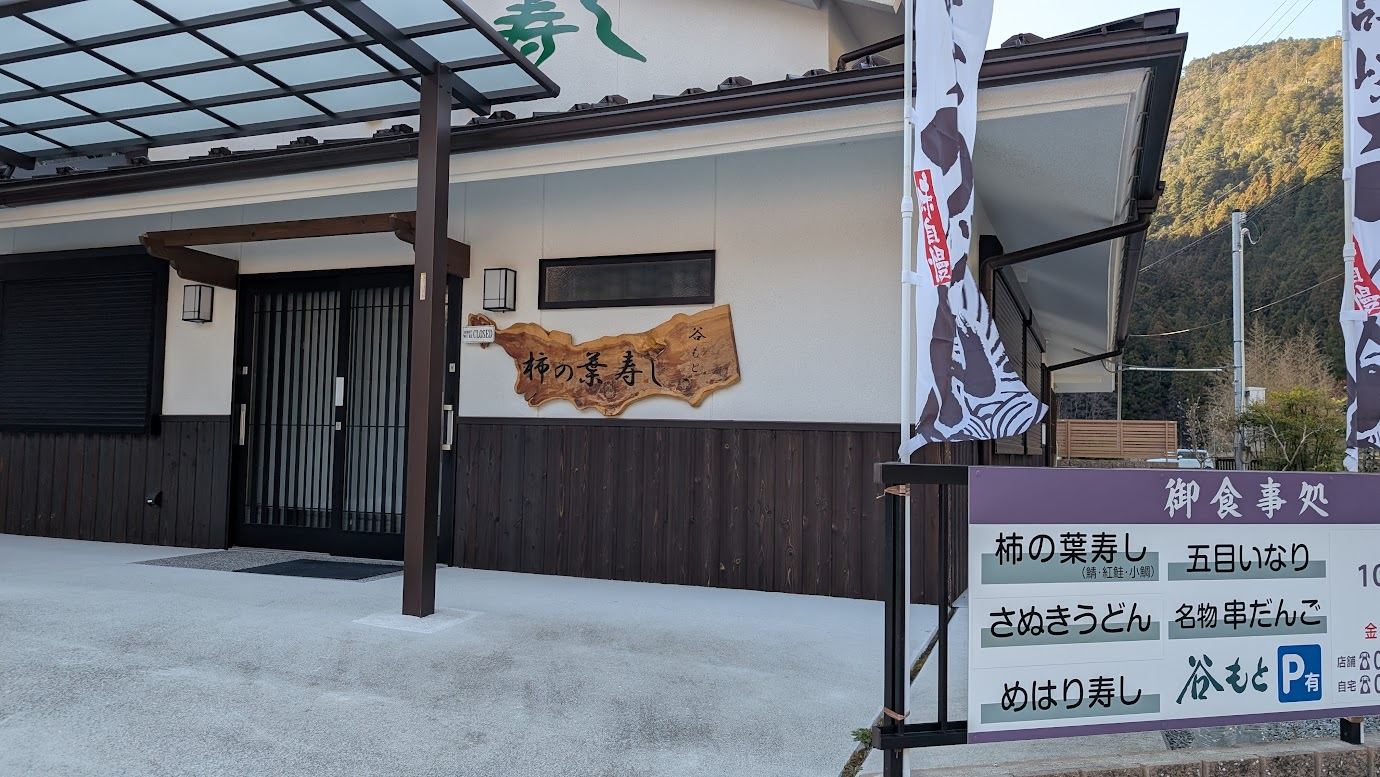 奈良県黒滝村、道の駅「吉野路 黒滝」の隣にある「柿の葉寿司 谷もと」の外観。木の看板に「柿の葉寿し 谷もと」と刻まれており、手前にはメニューの書かれた案内板が立っている。