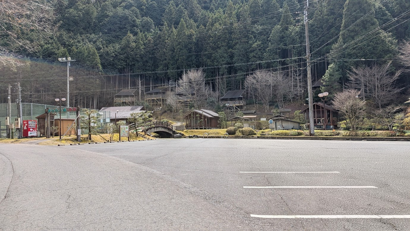 奈良県黒滝村にあるレジャー施設「きららの森・赤岩」の駐車場。広い駐車スペースの奥に、木造のコテージや小さな太鼓橋、管理棟が見える。背景には鬱蒼とした杉林が広がっている。