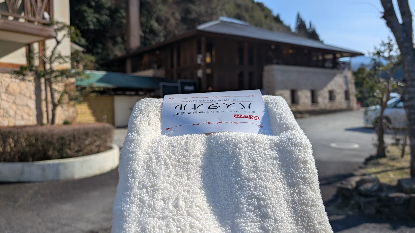 奈良県黒滝村にある温泉施設「黒滝の湯」を背景に、手前に真っ白なバスタオルを掲げた写真。背景には情緒ある木造の建物が見える。
