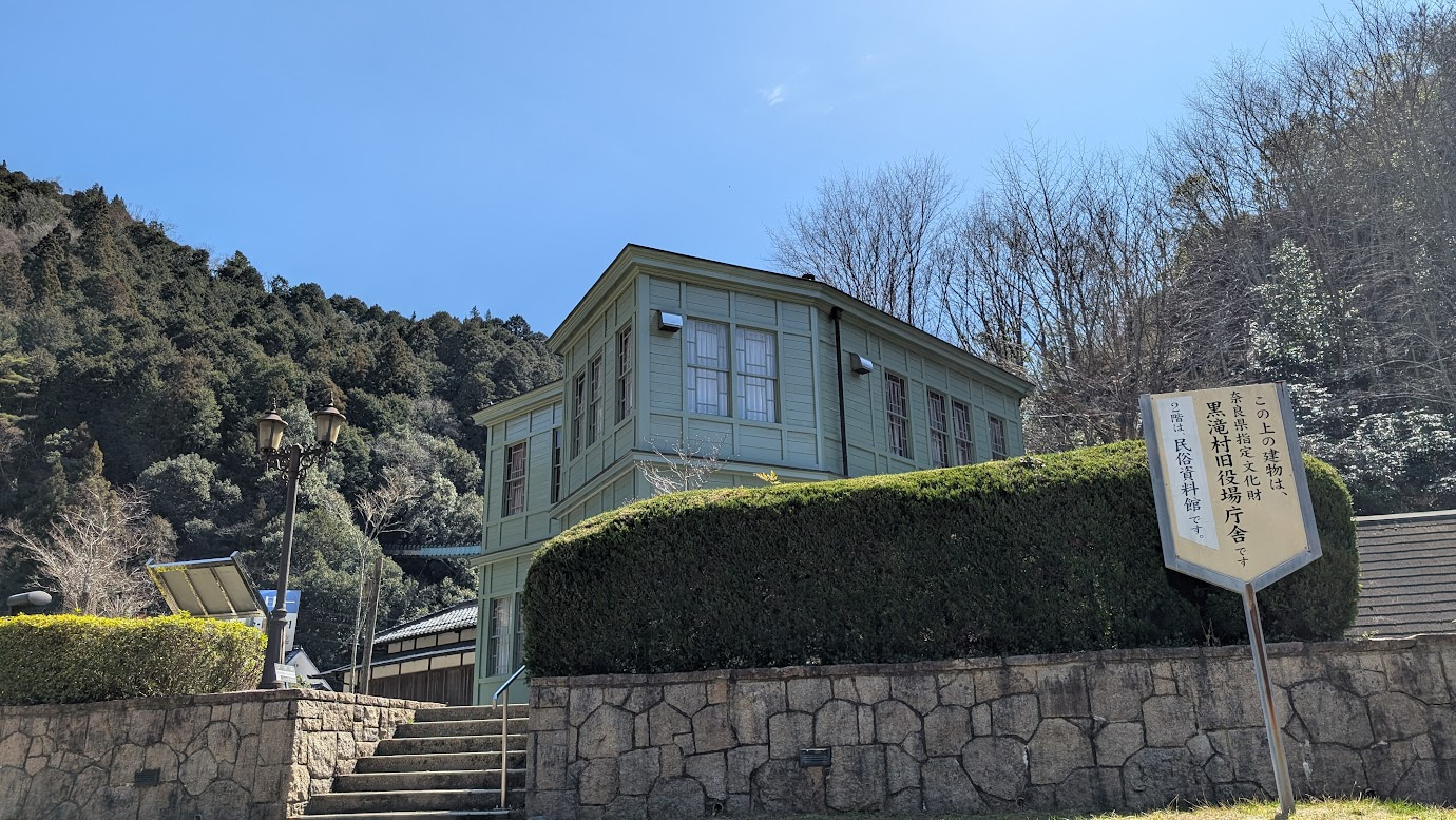 奈良県黒滝村にある旧役場庁舎を近くから見上げた写真。淡いグリーンの木造2階建て洋館。手前には「奈良県指定文化財 黒滝村旧役場庁舎」と書かれた看板と石垣の階段、レトロな街灯がある。