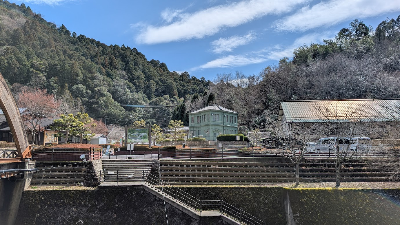 奈良県黒滝村にある淡いグリーンの木造2階建て洋風建築。黒滝村旧役場庁舎（現・文化保存伝習館）。手前には川と石積みの堤防があり、背後には深い山々が広がっている。