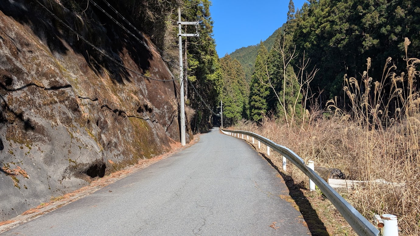 奈良県黒滝村の狭い村道。左側に巨大な岩壁、右側に白いガードレールと深い山々が広がる景色。青空の下、道が奥へと続いている。