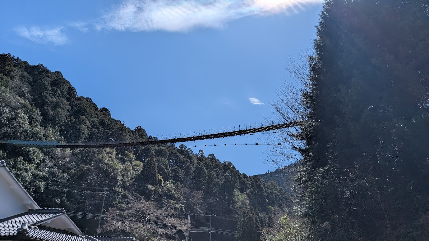 奈良県黒滝村にある黒滝吊橋を真下から見上げた写真。青空を背景に、細長い吊り橋が左右の山の斜面を繋いでいる。手前には和風建築の屋根が一部写っている。